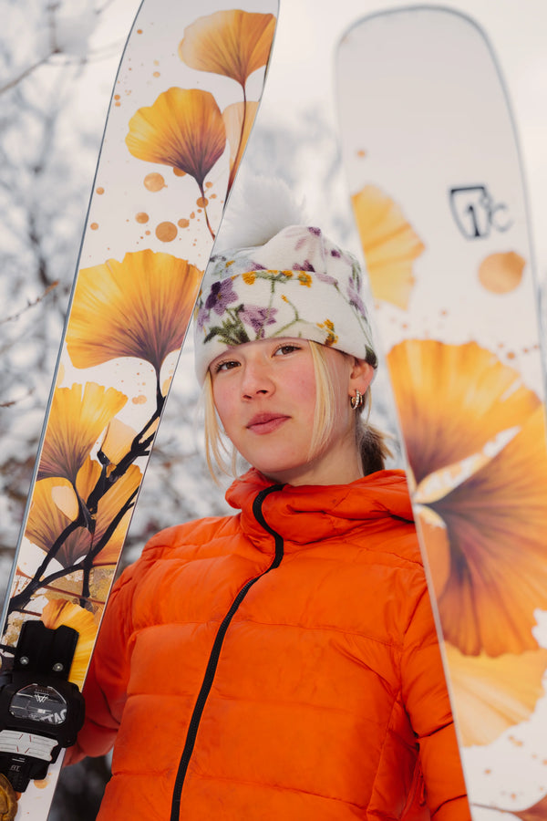 A person in an orange jacket and floral hat holds Icelantic Skis’ Tempest 94 skis with yellow flower designs, standing outdoors in a snowy setting.