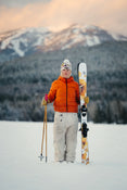Clad in an orange jacket and white pants, a person stands in the snow holding Icelantic Skis’ Tempest 94 ski poles and skis, with snowy mountains and trees stretching into the pastel sky behind them.