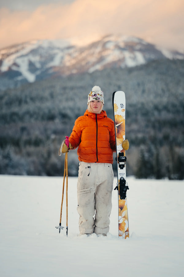 Clad in an orange jacket and white pants, a person stands in the snow holding Icelantic Skis’ Tempest 94 ski poles and skis, with snowy mountains and trees stretching into the pastel sky behind them.