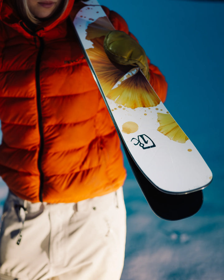 Wearing an orange jacket, white pants, and green gloves, a person stands on snow holding Icelantic Skis’ Tempest 94 skis—white with a yellow pattern—their face partially out of frame.