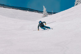 Wearing blue gear and a helmet, a skier carves down a snowy slope on a sunny day using Icelantic Skis’ Torrent 88, with a snow-covered tree and fence visible in the background.