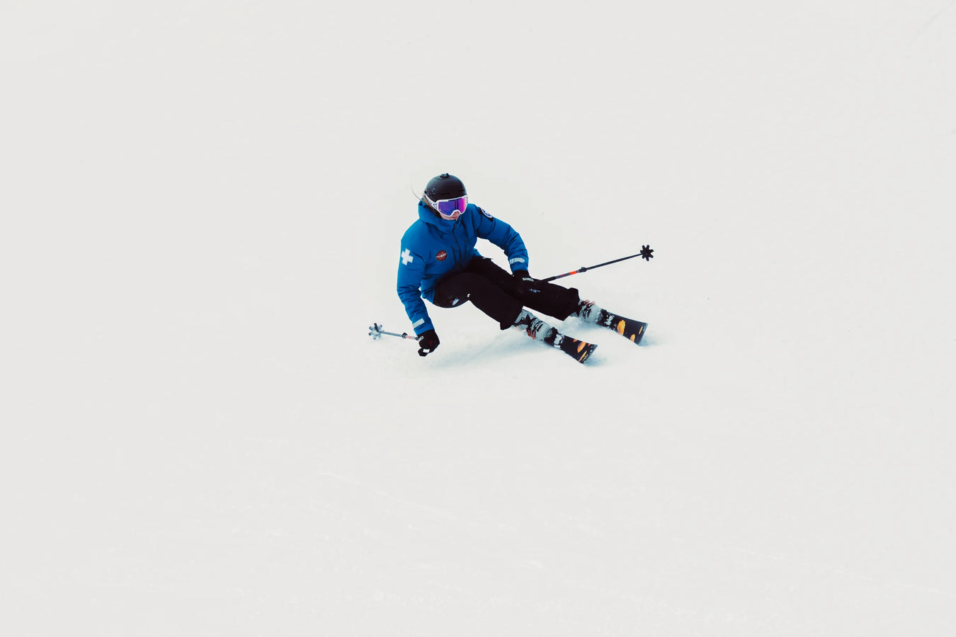 A skier in a blue jacket, black pants, and helmet carves through fresh snow on Icelantic Skis’ Tempest 88, holding ski poles out to the sides against a white, snowy backdrop with no visible landmarks.