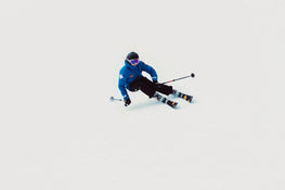 A skier in a blue jacket, black pants, and helmet carves through fresh snow on Icelantic Skis’ Tempest 88, holding ski poles out to the sides against a white, snowy backdrop with no visible landmarks.