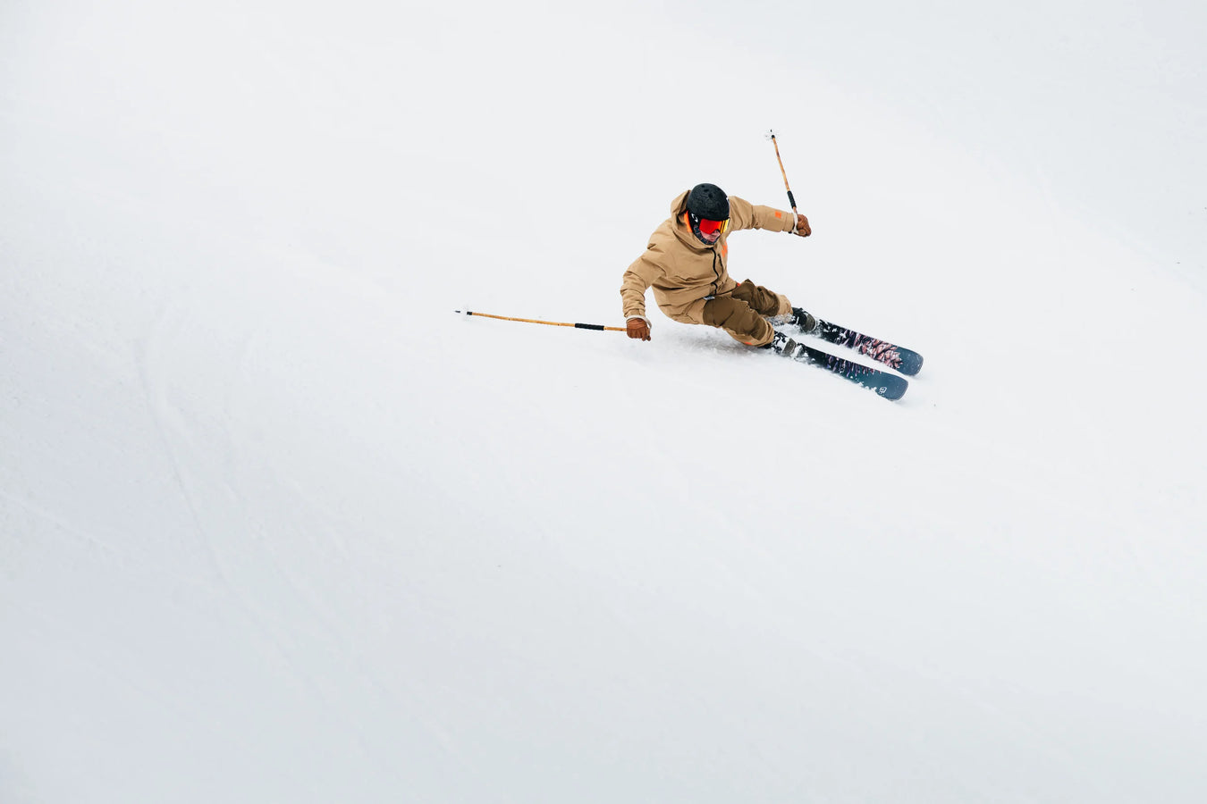 Wearing a tan jacket, black helmet, and red goggles, a skier carves down a snowy slope on Icelantic Skis’ Torrent 102, using ski poles for balance against the white, wintry background.