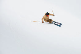 Wearing a tan jacket, black helmet, and red goggles, a skier carves down a snowy slope on Icelantic Skis’ Torrent 102, using ski poles for balance against the white, wintry background.
