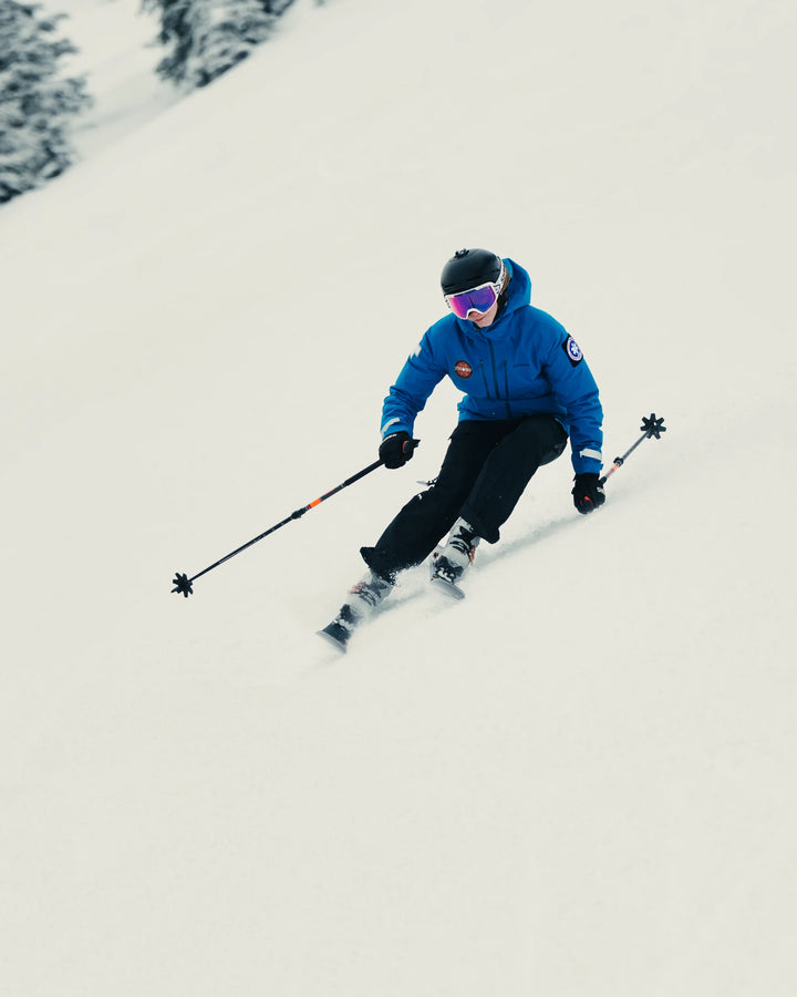A skier wearing a blue jacket, black pants, helmet, and goggles carves turns down a snowy slope on the Icelantic Skis Tempest 88, gripping ski poles as snow-covered trees stand in the background.