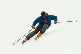 A skier in a blue jacket, gray pants, and orange boots makes a sharp turn on a snowy slope using the Icelantic Skis Torrent 96 for balance. The skier wears a black helmet and reflective goggles.