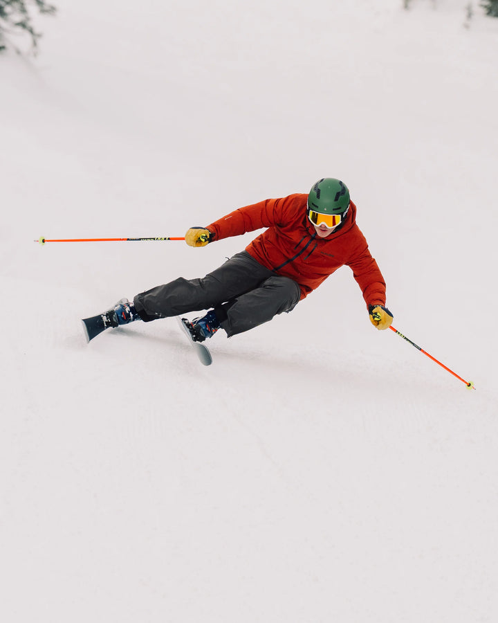 A skier in a red jacket, black pants, and green helmet carves through fresh snow on a snowy slope, leaning sharply with ski poles extended while riding the Torrent 88 by Icelantic Skis.