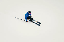 A skier in a blue jacket and black helmet carves through fresh snow on Icelantic Skis Tempest 88, gripping ski poles while making a turn.