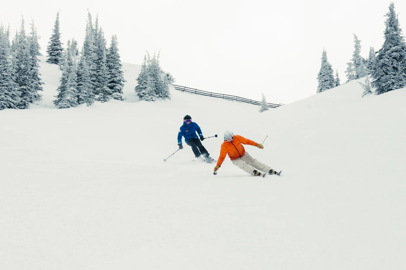 Two people carve down a snowy slope on their Icelantic Skis Tempest 94, weaving between snow-covered trees—one skier in an orange jacket leading the way, closely followed by another in a blue jacket.
