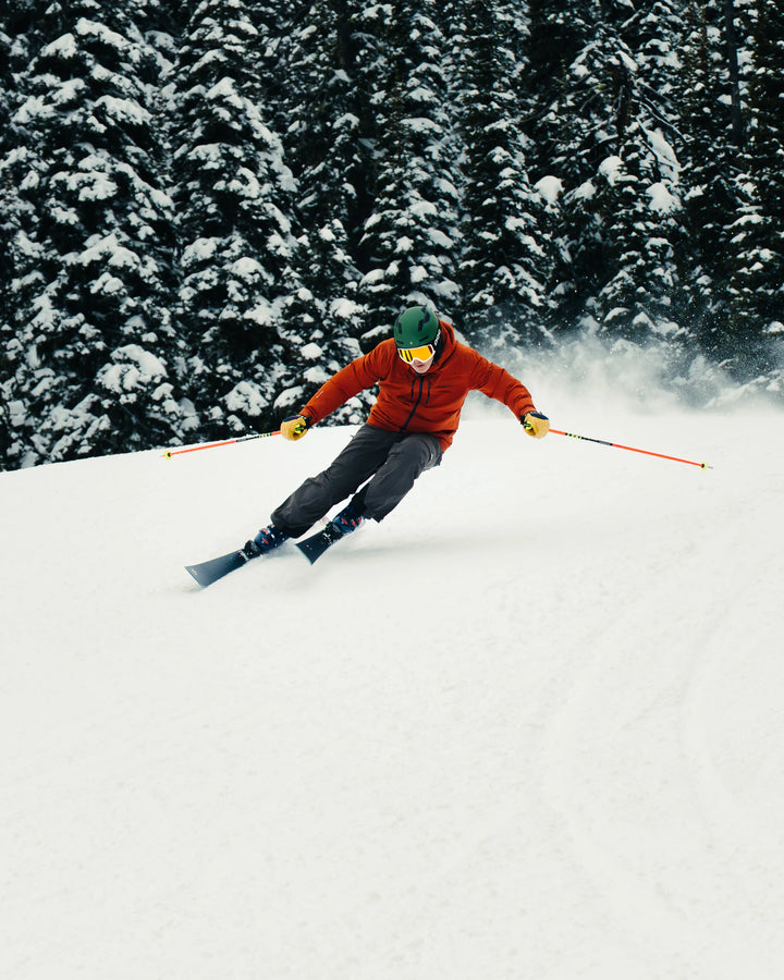 Wearing a red jacket and helmet, a skier carves down a snowy slope lined with snow-covered pines on their Icelantic Skis Torrent 88.