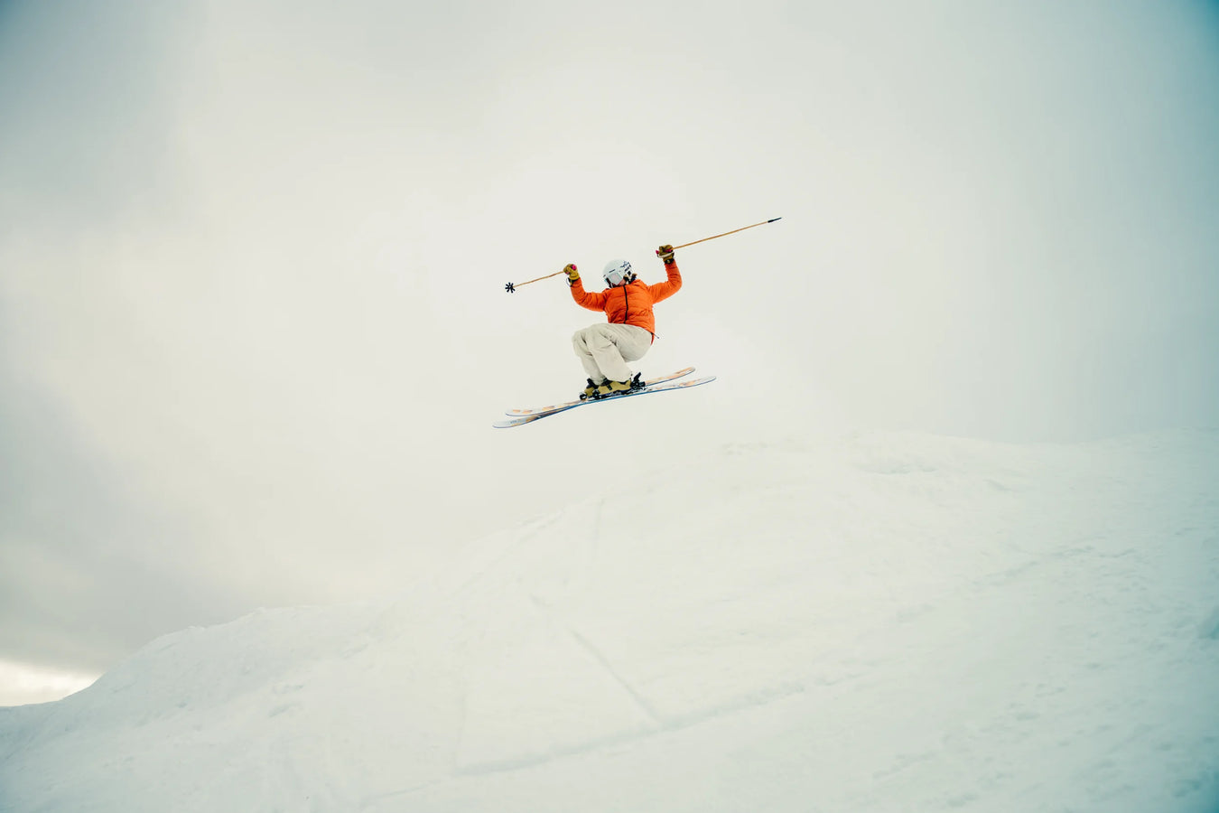 Soaring high above a snowy slope under a cloudy sky, a skier in an orange jacket and white pants grips their ski poles while catching air on their Icelantic Skis Tempest 94.