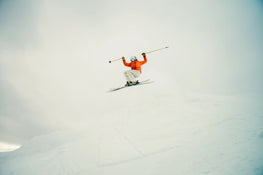 Soaring high above a snowy slope under a cloudy sky, a skier in an orange jacket and white pants grips their ski poles while catching air on their Icelantic Skis Tempest 94.