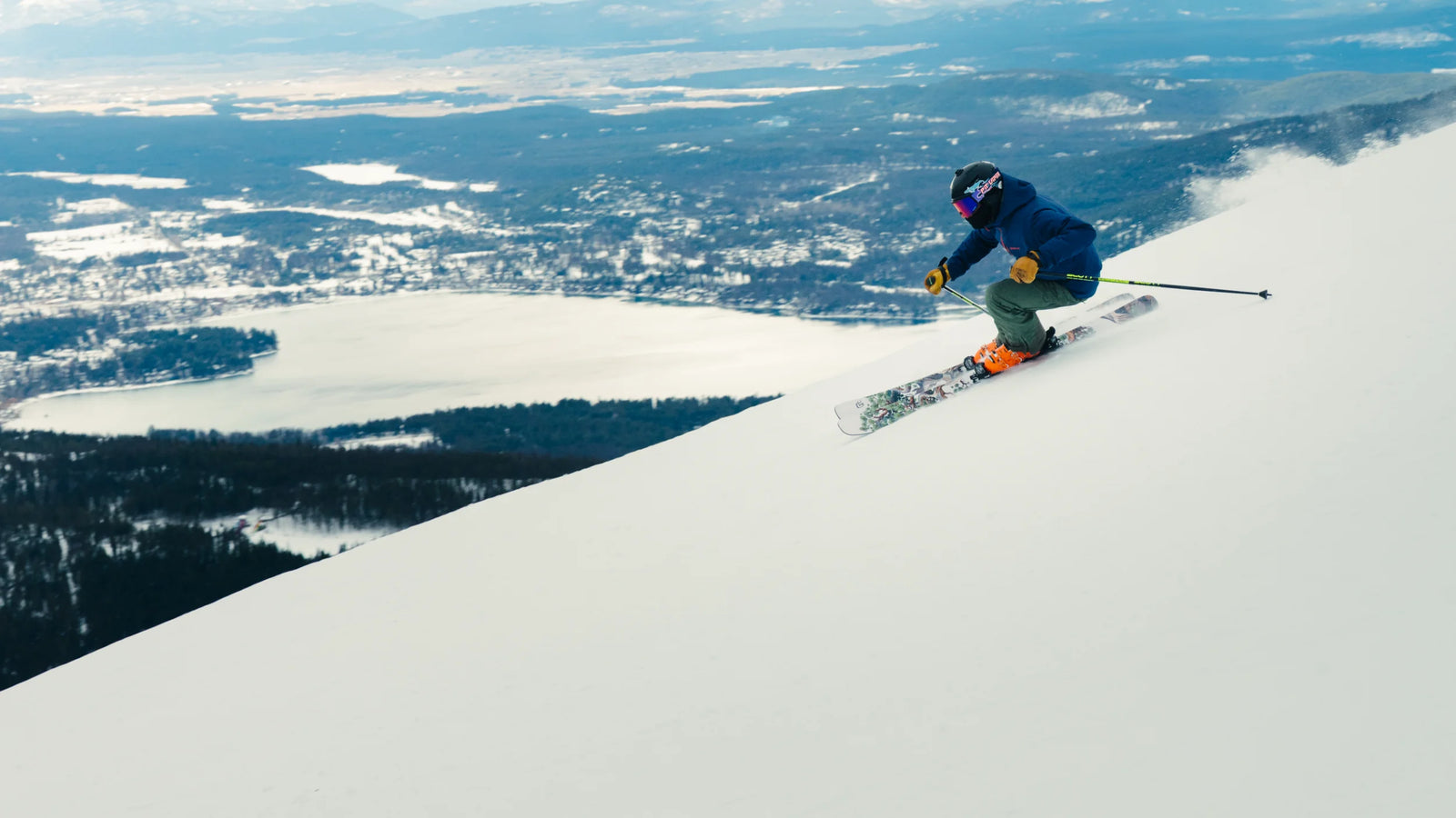 A skier on Icelantic Skis’ Torrent 96, dressed in blue and green gear, speeds down a snowy slope with a lake, forest, and distant mountains in the background beneath a cloudy sky.