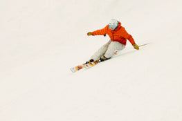 An athlete wearing an orange jacket and white helmet carves downhill on a snowy slope atop the Icelantic Skis Tempest 94, leaning into a turn with both arms outstretched for balance.