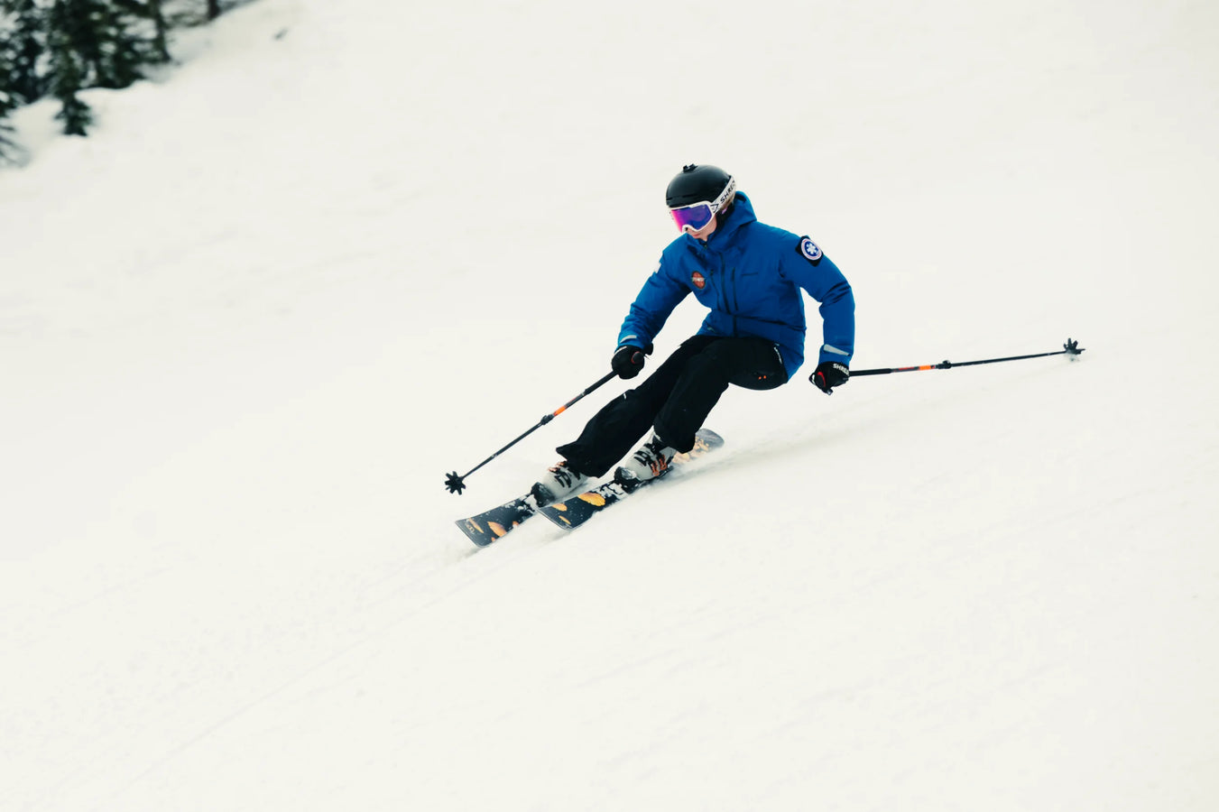 A skier equipped with Icelantic Skis’ Tempest 88 glides down a snowy slope in a blue jacket and black helmet, leaning into a turn with ski poles in hand. The mostly white background features a hint of trees in the upper left.