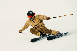 A skier equipped with the Icelantic Skis Torrent 102 in tan gear and a black helmet carves through snow, leaning forward with ski poles extended and knees bent, creating dynamic motion on a snowy slope.
