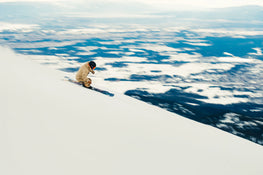 Wearing beige gear and a black helmet, a skier carves down a steep, snow-covered slope on Icelantic Skis’ Torrent 102, with a blur of blue and white mountains forming the backdrop.