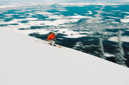 A skier in an orange jacket and helmet carves down a steep, snowy slope on Icelantic Skis’ Tempest 94, with a blurred landscape of snow and trees in the background.