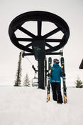 Clad in blue winter gear, a skier stands in the snow holding the Icelantic Skis Tempest 88, positioned beneath a large ski lift wheel with empty chairs. Snow-covered trees and a cloudy sky can be seen in the background.