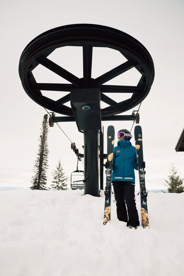 Clad in blue winter gear, a skier stands in the snow holding the Icelantic Skis Tempest 88, positioned beneath a large ski lift wheel with empty chairs. Snow-covered trees and a cloudy sky can be seen in the background.