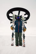 Clad in winter gear, a skier holds two Icelantic Skis Torrent 96 skis while standing before a circular ski lift station amid snowy conditions.