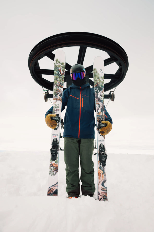 Clad in winter gear, a skier holds two Icelantic Skis Torrent 96 skis while standing before a circular ski lift station amid snowy conditions.