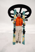 A skier in an orange jacket and white pants stands in the snow, holding green Icelantic Skis Tempest 100 all-mountain skis with yellow flower designs featuring a poplar and birch wood core, with a ski lift wheel in the background under a cloudy sky.