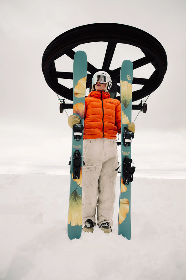 A skier in an orange jacket and white pants stands in the snow, holding green Icelantic Skis Tempest 100 all-mountain skis with yellow flower designs featuring a poplar and birch wood core, with a ski lift wheel in the background under a cloudy sky.