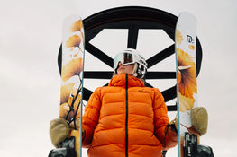 A person in an orange jacket and ski helmet stands holding Icelantic Skis’ Tempest 94 skis, featuring orange floral designs, with a ski lift wheel overhead against a cloudy sky.