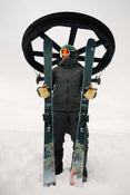 A skier in black gear and orange goggles stands in the snow, holding a pair of Icelantic Skis Torrent 88 upright, with a ski lift wheel visible overhead against a cloudy sky.