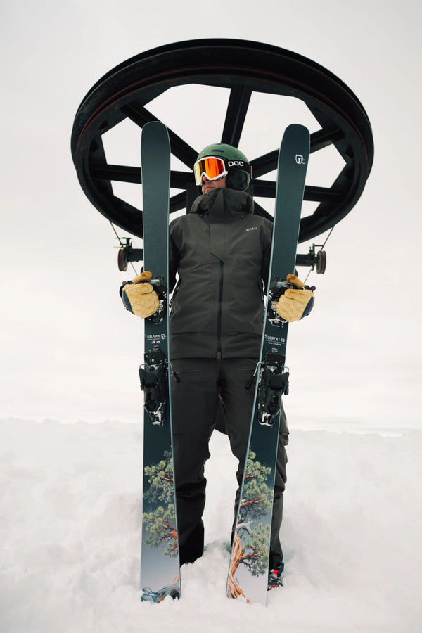 A skier in black gear and orange goggles stands in the snow, holding a pair of Icelantic Skis Torrent 88 upright, with a ski lift wheel visible overhead against a cloudy sky.