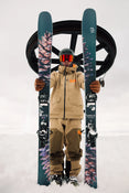 A skier in tan gear and orange goggles stands in the snow, holding a pair of Icelantic Skis Torrent 102 upright. Overhead, a ski lift wheel is visible against the cloudy sky.