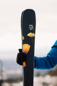 A person in a blue jacket and black glove holds an Icelantic Skis Tempest 88, a dark ski decorated with yellow-orange flowers and a logo. The snowy background is blurred.