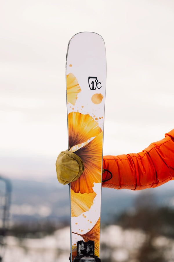 A person in an orange jacket holds up the Icelantic Skis Tempest 94, featuring a white base with an orange-yellow floral design, against a snowy outdoor background.
