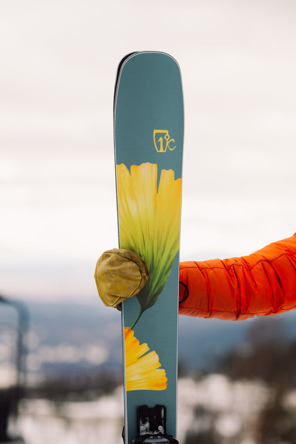 A person in an orange jacket holds the Icelantic Skis Tempest 100, an all-mountain ski featuring a yellow flower design and a high-performance poplar and birch wood core with carbon matrix; a snowy, blurred landscape forms the background.