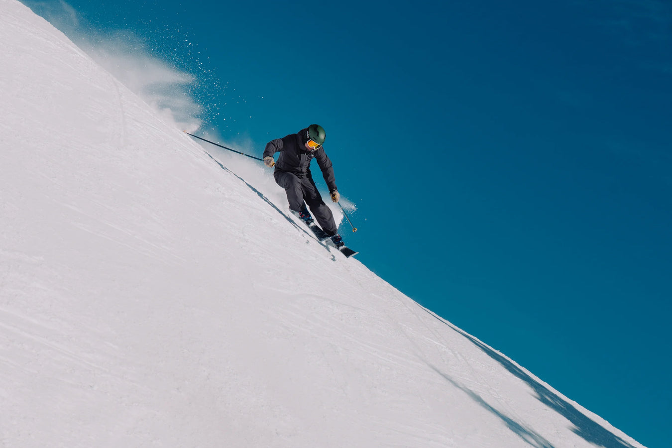 Clad in dark gear and a green helmet, a skier on Icelantic Skis’ Torrent 88 carves down a steep, snowy slope beneath a clear blue sky, spraying powder in their wake.