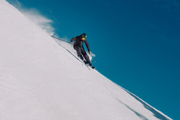 Clad in dark gear and a green helmet, a skier on Icelantic Skis’ Torrent 88 carves down a steep, snowy slope beneath a clear blue sky, spraying powder in their wake.