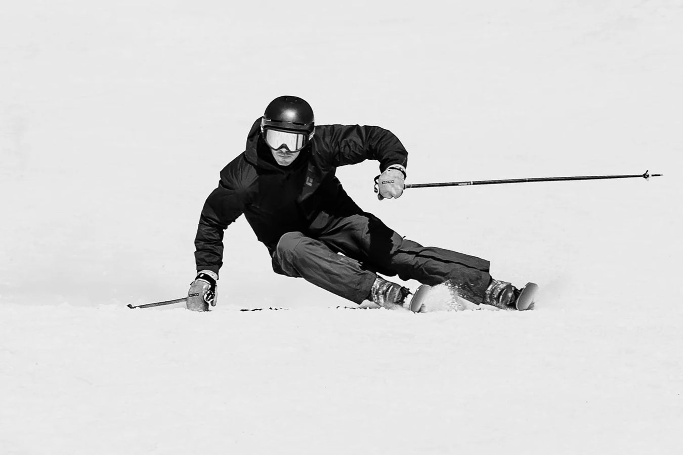 Wearing dark gear and a helmet, a skier carves a sharp turn on the snowy slope, leaning low with both poles angled outward on their Icelantic Skis Torrent 88.