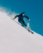 An adventurous skier in blue gear and helmet carves a sharp turn on a snowy slope, sending up powder as they ride the Icelantic Skis Torrent 96 beneath a clear blue sky.