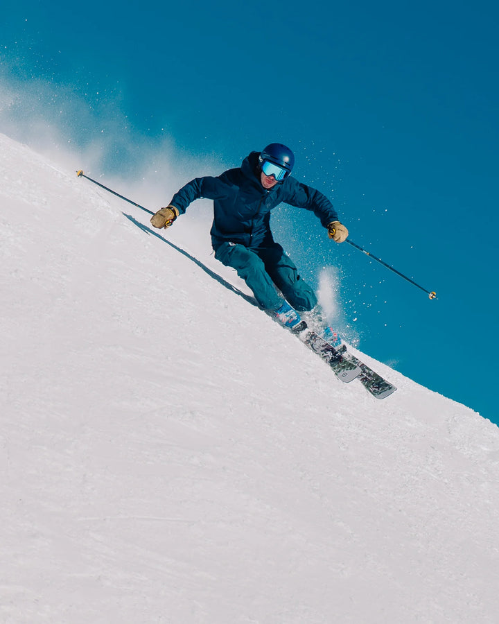 An adventurous skier in blue gear and helmet carves a sharp turn on a snowy slope, sending up powder as they ride the Icelantic Skis Torrent 96 beneath a clear blue sky.