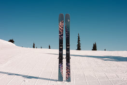 A pair of Icelantic Skis Torrent 102, featuring a distinctive pinecone design, stands upright in the snow on a groomed ski slope, with pine trees and a clear blue sky in the background.