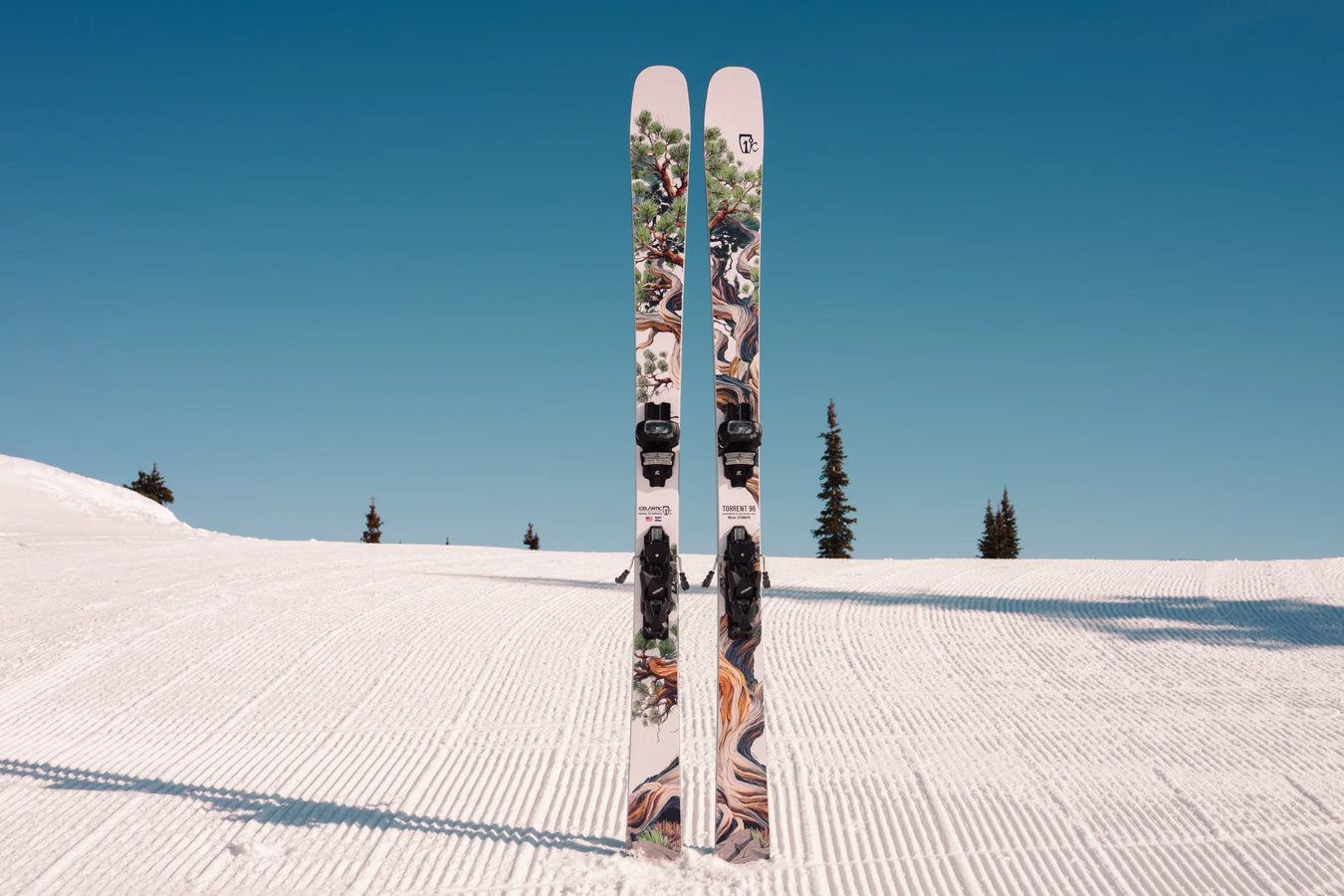 A pair of Icelantic Skis Torrent 96, featuring colorful patterns, stands upright in the snow on a groomed ski slope beneath a clear blue sky, with a few pine trees in the background.