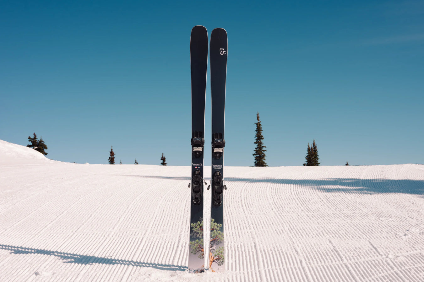 A pair of Icelantic Skis Torrent 88 stand upright on a freshly groomed, snow-covered slope beneath a clear blue sky, with a few trees in the background.