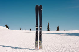 A pair of Icelantic Skis Torrent 88 stand upright on a freshly groomed, snow-covered slope beneath a clear blue sky, with a few trees in the background.