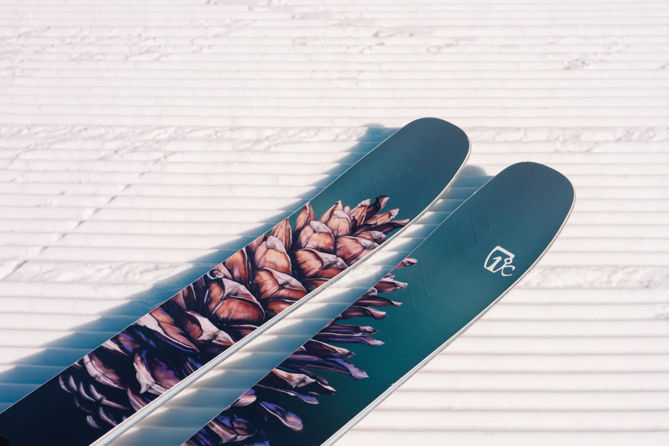 A close-up of two Icelantic Skis Torrent 102 skis, featuring a pine cone design, resting on freshly groomed, parallel-lined snow.