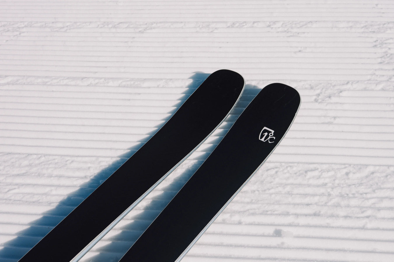 A close-up of two Icelantic Skis Torrent 88 black skis, featuring a white logo, resting on freshly groomed, corduroy-patterned snow under bright sunlight.