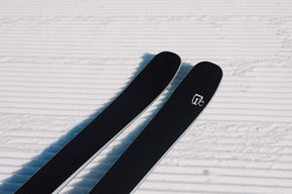 A close-up of two Icelantic Skis Torrent 88 black skis, featuring a white logo, resting on freshly groomed, corduroy-patterned snow under bright sunlight.