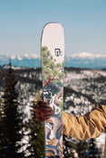 A person in a tan jacket and brown glove holds the Icelantic Skis Torrent 96, which features a colorful nature-themed design; snowy mountains and pine trees are visible in the background under a clear sky.