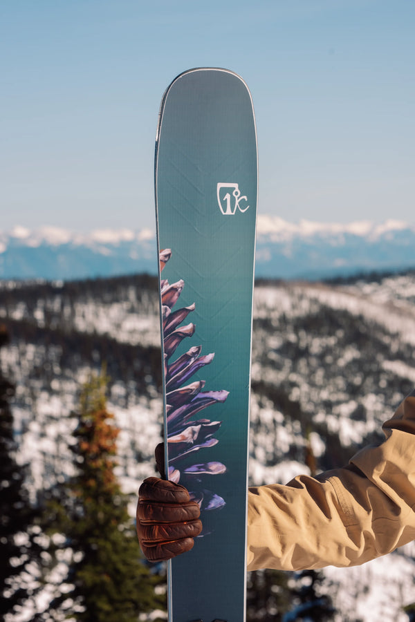 A person in a tan jacket and brown glove holds the Torrent 102 ski by Icelantic Skis, featuring a floral or plant design and a white logo near the top, with a mountain and forest landscape in the snowy background.